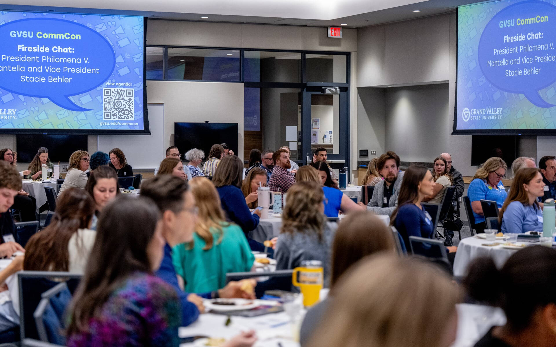 at GVSU CommCon, people seated at round tables, screens show QR code to an agenda and word bubble with Fireside Chat: President Mantella and Vice President Stacie Behler.